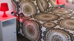 Une chambre à coucher avec une couvre-lit à motifs, deux tables de chevet avec des lampes rouges, un tableau représentant un coucher de soleil accroché au mur, et une fenêtre avec des rideaux.