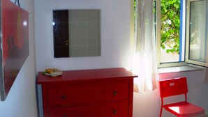 Une pièce avec une commode rouge, un miroir, un climatiseur, une fenêtre donnant sur un paysage verdoyant et un fauteuil rouge.