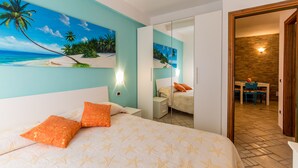 Een slaapkamer met een bed, oranje kussens, een schilderij van een strand, een spiegel en uitzicht op een eetgedeelte.