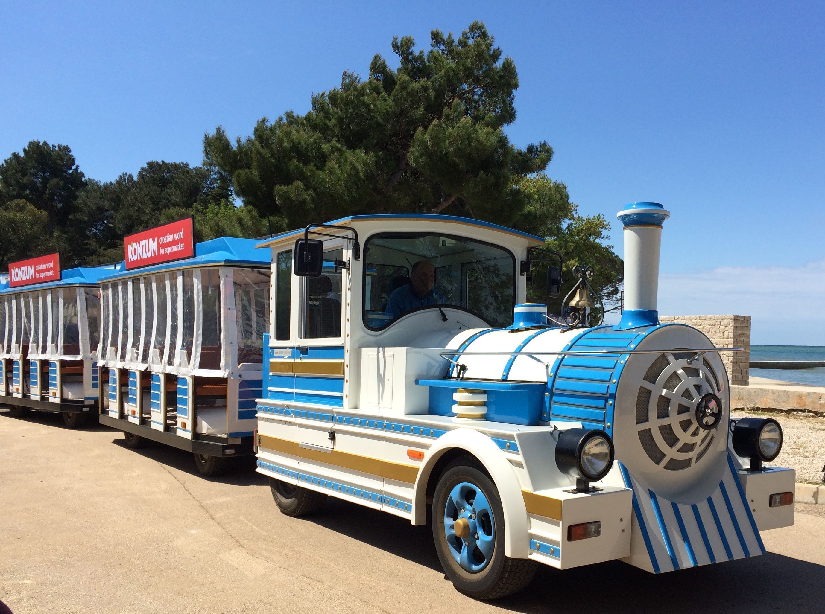 Ein blau-weiß gestreifter Zug mit Café-Wagen, geparkt auf einem Betonweg. Im Hintergrund Bäume und ein Gewässer.
