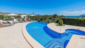 Ein gewundener Pool mit Liegestühlen und Sonnenschirmen, umgeben von Grünpflanzen und mit Blick auf das Meer und die Hügel.
