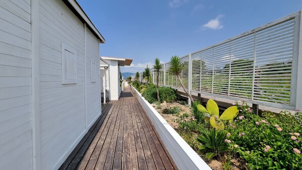 A narrow wooden pathway flanked by white buildings and a garden with various plants and flowers.