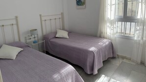 A bedroom with two single beds, a nightstand with a lamp, a window with curtains, and a wall-mounted peacock painting.
