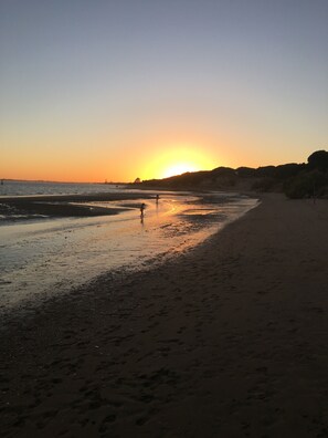 Une plage au coucher du soleil, avec des empreintes de pas et quelques personnes qui se promènent.
