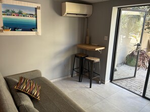 Ein modernes Wohnzimmer mit einem grauen Sofa, einem bunten Kissen, einem kleinen Holztisch und zwei Hockern, sowie einem großen Fenster mit Blick ins Freie.