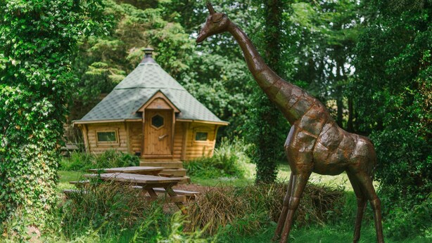 A wooden cabin with a thatched roof surrounded by lush greenery and a giraffe statue. - Corffe Cottages, Space for all the family, heated indoor pool (Barnstaple)