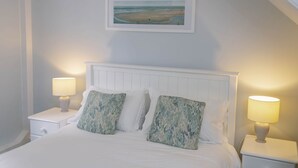 A neatly made bed with white linens and two decorative pillows, flanked by two bedside tables with lamps, in a room with a white headboard and a framed picture on the wall. - Corffe Cottages, Space for all the family, heated indoor pool (Barnstaple)