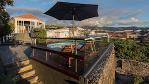 Area piscina sul tetto con ringhiera in vetro, pavimento in legno e un ombrellone grande.