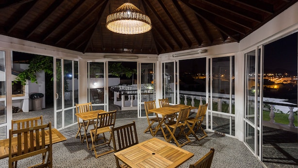 Un'area pranzo all'aperto riparata, con tavoli e sedie in legno, ampie finestre e un lampadario.