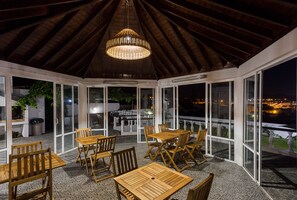 A covered outdoor dining area with wooden tables and chairs, large windows, and a hanging light fixture. - Quinta das Rosas Villas - White Rose (Ribeira Grande)