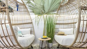A room with two wicker chairs, a small round table with a bowl of fruit, and a tall plant.