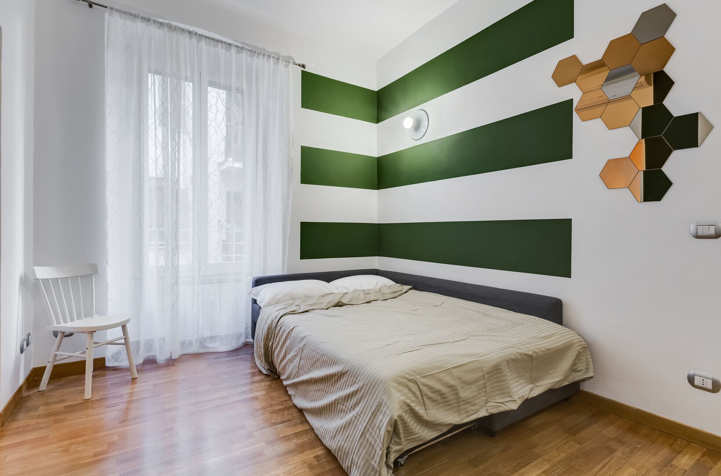 A bedroom with a bed, a chair, a window with curtains, and a wall with geometric patterns.