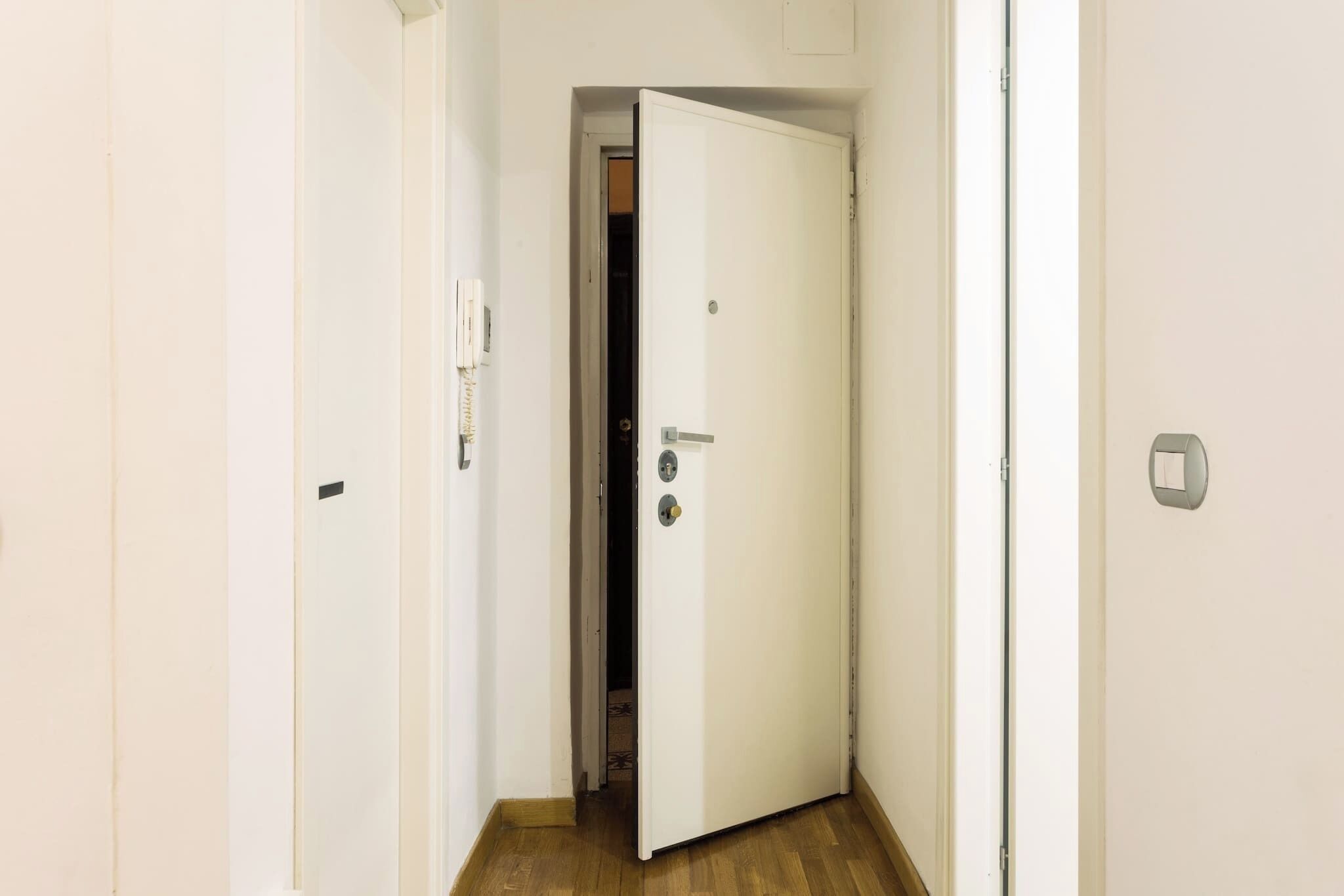 Un pasillo estrecho con una puerta blanca al final, un interruptor de luz en la pared y suelo de madera.