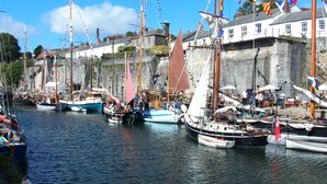 Un puerto con varios veleros amarrados a lo largo de la orilla, rodeado de edificios de piedra y banderas.