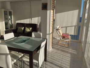 Un comedor con mesa de vidrio, sillas blancas, un sofá de cuero marrón con cojines estampados y un balcón soleado con una silla.