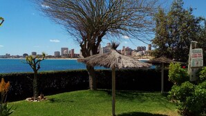 A grassy area with a tree and a thatched structure, a paved walkway, and a distant cityscape. - PRIVATE ACCESS TO BEACH IN THE SAME URBANIZATION (El Campello)