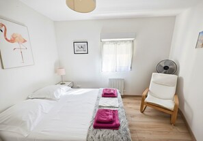 A bedroom with a bed, a chair, a radiator, a window with curtains, and a wall-mounted fan. - Cottage with Pool, Fenced Garden – Pets Welcome (Saint-Gilles)