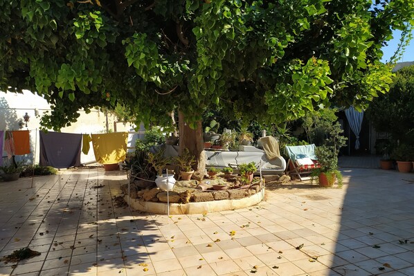 A courtyard with a tiled floor, potted plants, a tree providing shade, and a small seating area.