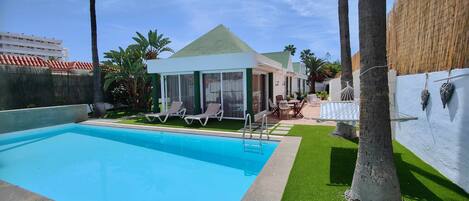 A swimming pool with lounge chairs, a covered patio, and a view of a building with a green roof.