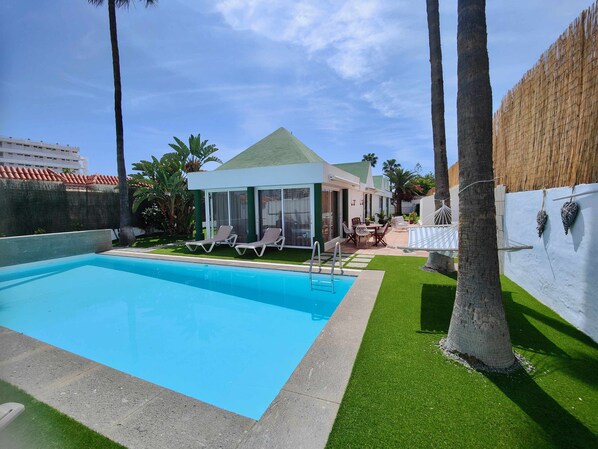 A swimming pool with lounge chairs, a covered patio, and a view of a building with a green roof.