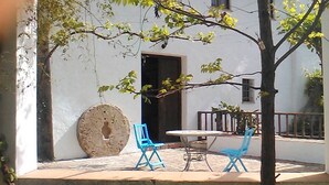 Um pátio com uma mesa e duas cadeiras azuis, uma pedra decorativa grande e uma parede branca com uma janela.