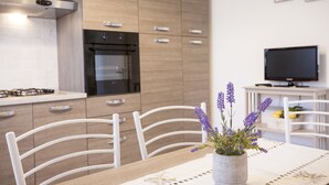 A modern kitchen with wooden cabinets, a built-in oven, and a dining area with white chairs and a table. - APPARTAMENTI MARCIANA MARINA - APPARTAMENTO L'EDERA (Marciana Marina)