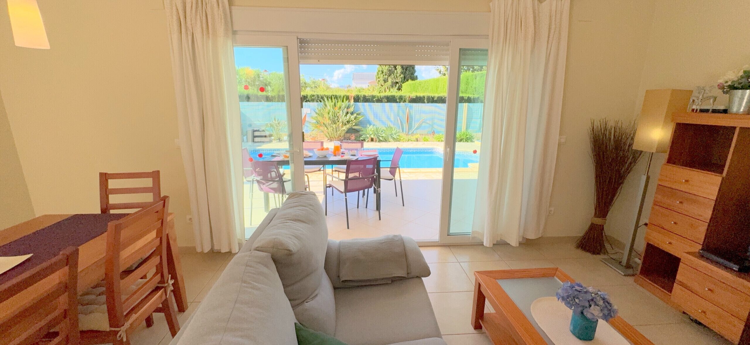 A living room with a sofa, a dining table with chairs, a TV stand, and a view of a pool and garden through a sliding glass door.