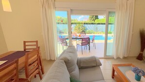 A living room with a sofa, a dining table with chairs, a TV stand, and a view of a pool and garden through a sliding glass door. - 0533 Villa Laura Poblets (Denia)
