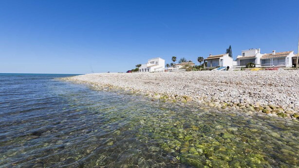 A pebble beach with clear water, white buildings, and palm trees. - 0533 Villa Laura Poblets (Denia)