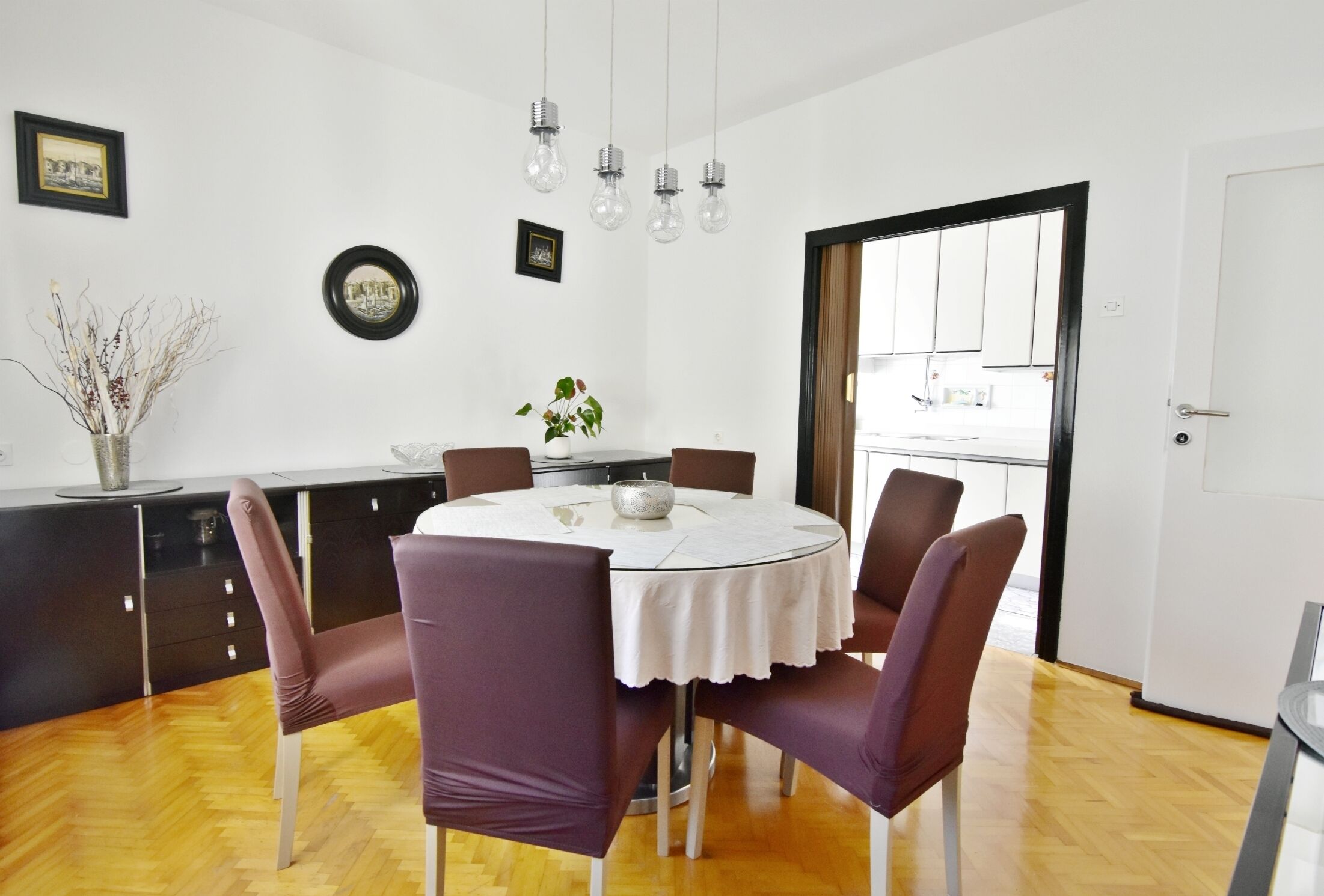 A dining area with a round table, six chairs, a vase with branches, and wall-mounted light fixtures.