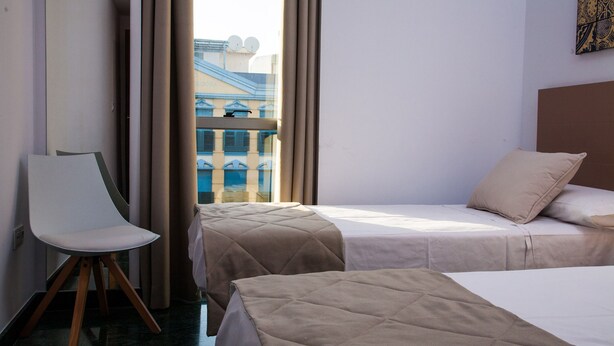 A hotel room with two beds, a chair, and a view of a building with blue windows. - Great penthouse in the center of Córdoba (Córdoba)