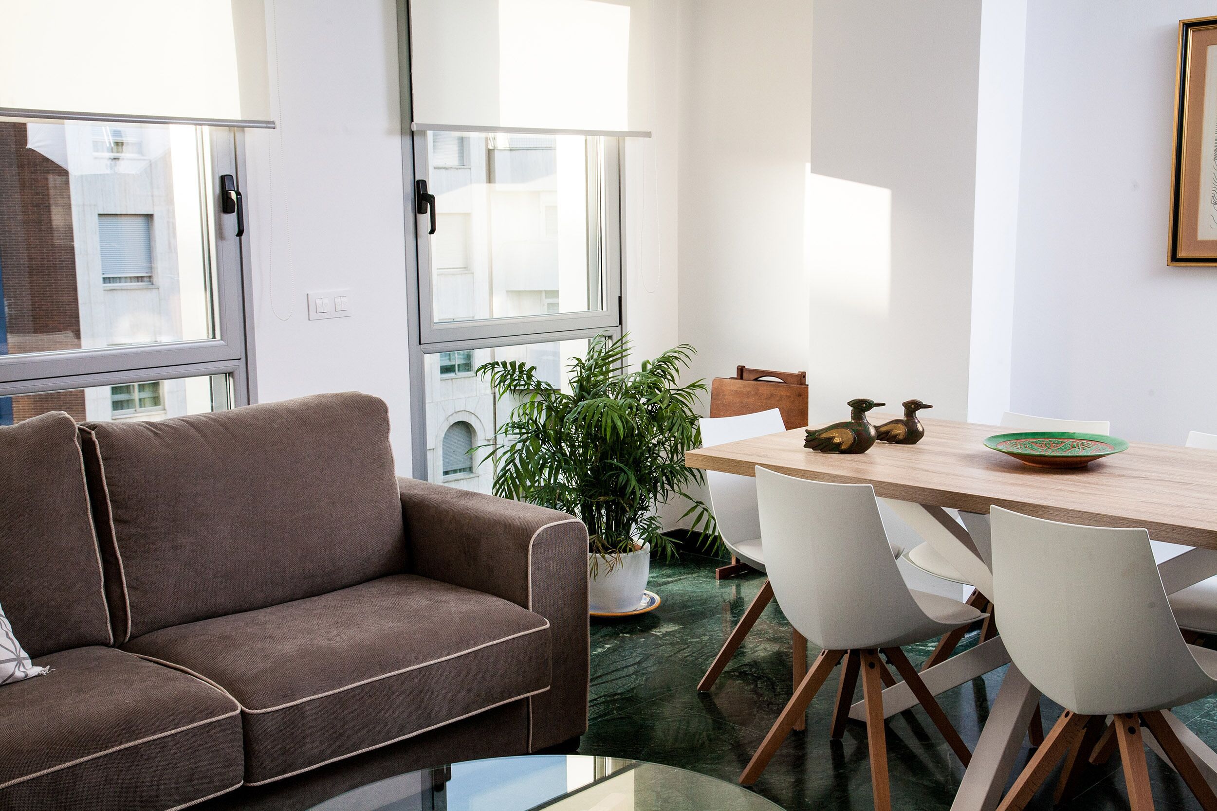 Een moderne woonkamer met een bruine bank, een eettafel met stoelen, een potplant en uitzicht buiten door grote ramen.