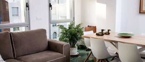 Een moderne woonkamer met een bruine bank, een eettafel met stoelen, een potplant en uitzicht buiten door grote ramen.