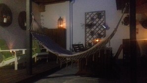 Una hamaca sobre una terraza de madera con barandilla, un farol colgante en la pared y una ventana con celosía.