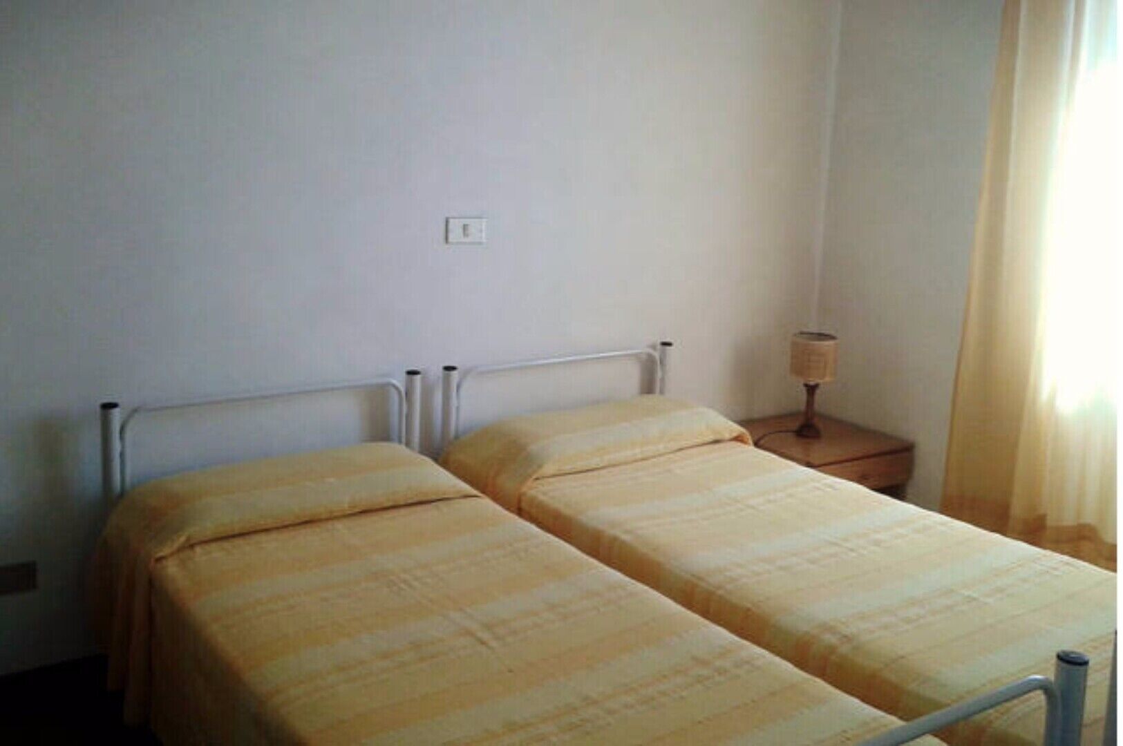 Een kleine, eenvoudige kamer met twee enkele bedden, een nachtkastje met lamp en een stopcontact aan de muur.