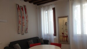 A living room with a dark sofa, a white round table, and red chairs. There are curtains, a mirror, and a door in the background.