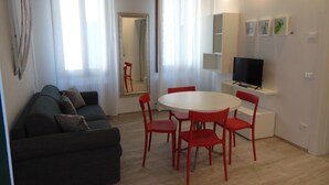 A modern living room with a grey sofa, red chairs, a white dining table, and a flat-screen TV on a white stand.