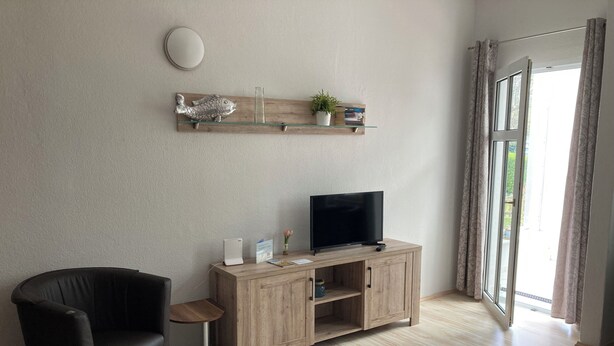 A living room with a wooden TV stand, a black leather chair, a small side table, and a large window with curtains. - Rosenbogen - Holiday apartment with conservatory and terrace (Wangerooge)