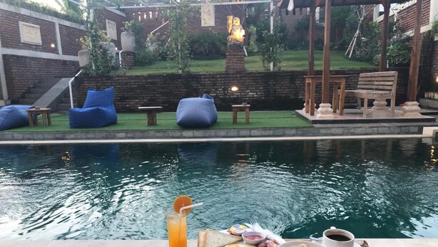 A poolside breakfast setup with a variety of food items and a drink, overlooking a pool and a garden area with seating and a statue. - Oka's Guest House (Canggu)