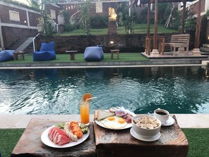 A poolside breakfast setup with a variety of food items and a drink, overlooking a pool and a garden area with seating and a statue. - Oka's Guest House (Canggu)