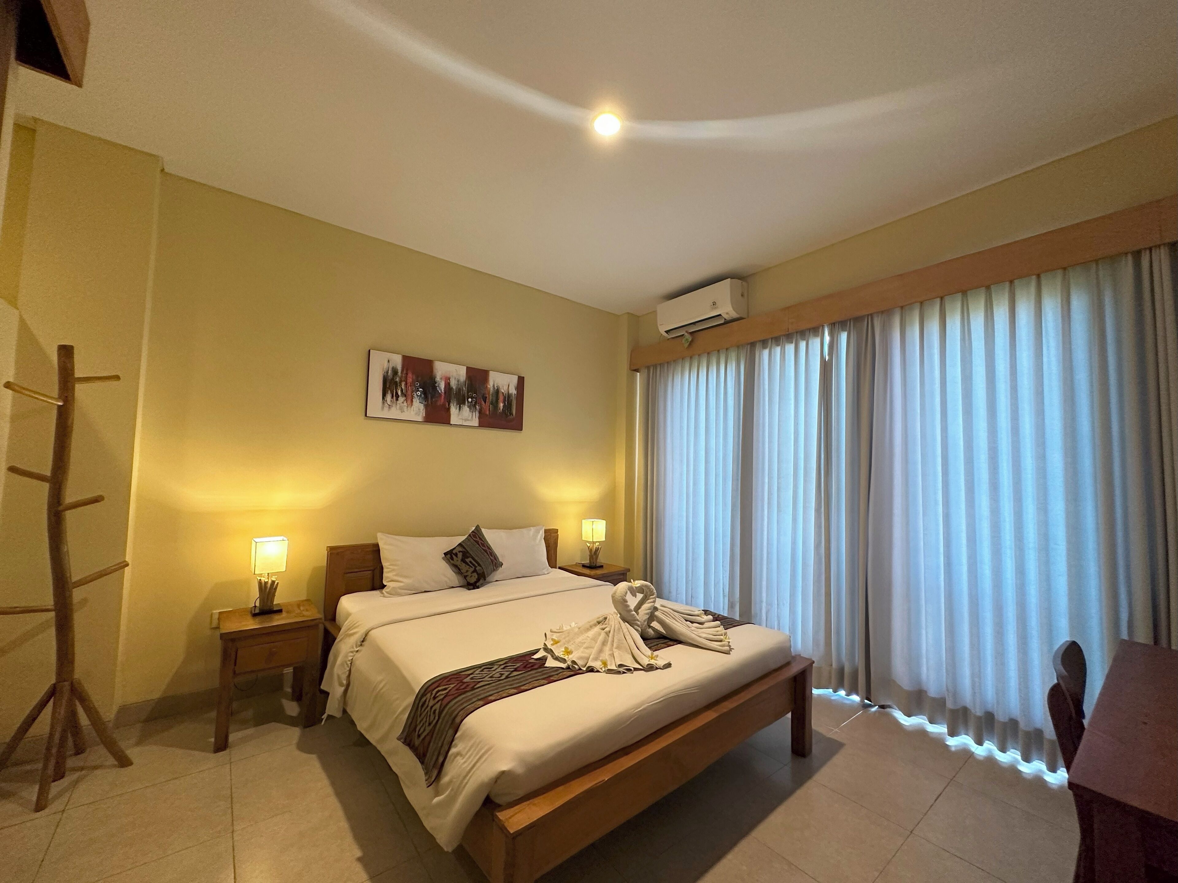 A hotel room with a bed, bedside lamps, a wall-mounted air conditioner, and a large window with curtains.
