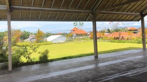 A covered outdoor area with a view of a green lawn and buildings in the background. - Oka's Guest House (Canggu)