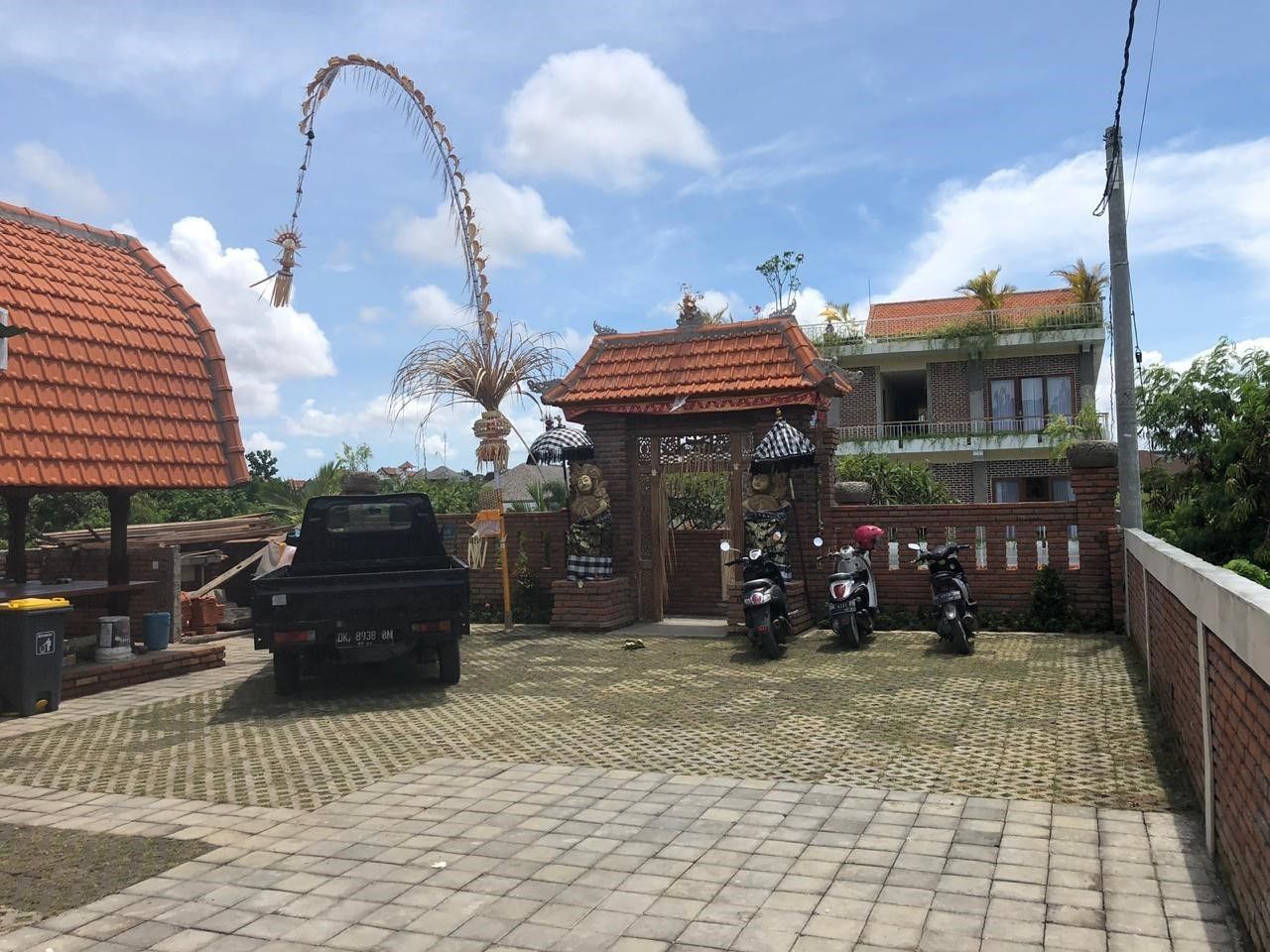 Une allée d’entrée pavée où sont garés un camion et des motos, un portail agrémenté d’éléments décoratifs et, au fond, un bâtiment avec un toit recouvert de tuiles rouges.