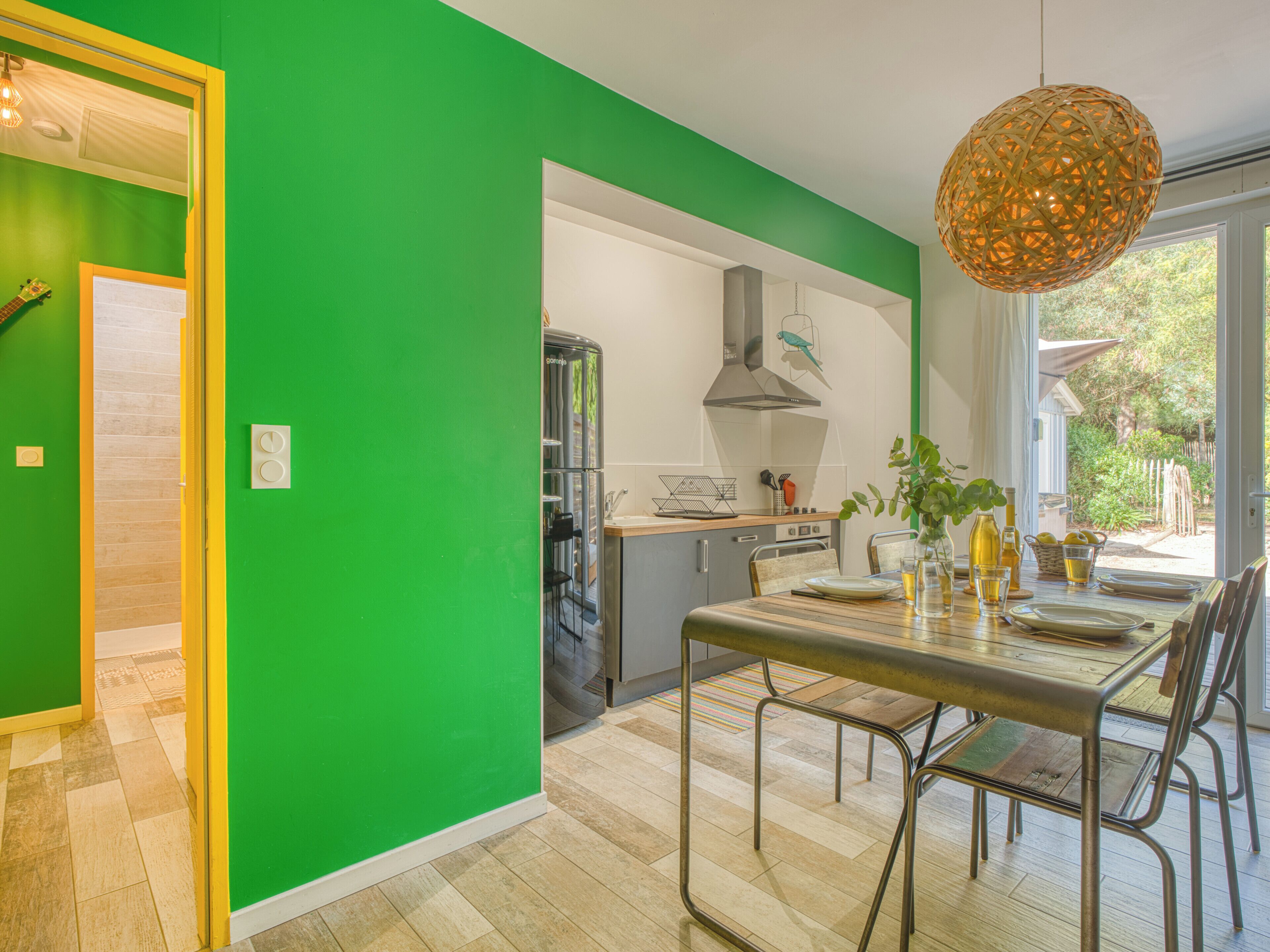 Une cuisine moderne avec un mur vert, un coin repas avec une table et des chaises, et une vue sur l’extérieur.
