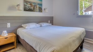 A bedroom with a bed, a nightstand, a wall-mounted lamp, a painting, and a window with a view. - Villa Brésil - Domaine Le Sherwood (Notre-Dame-De-Monts)