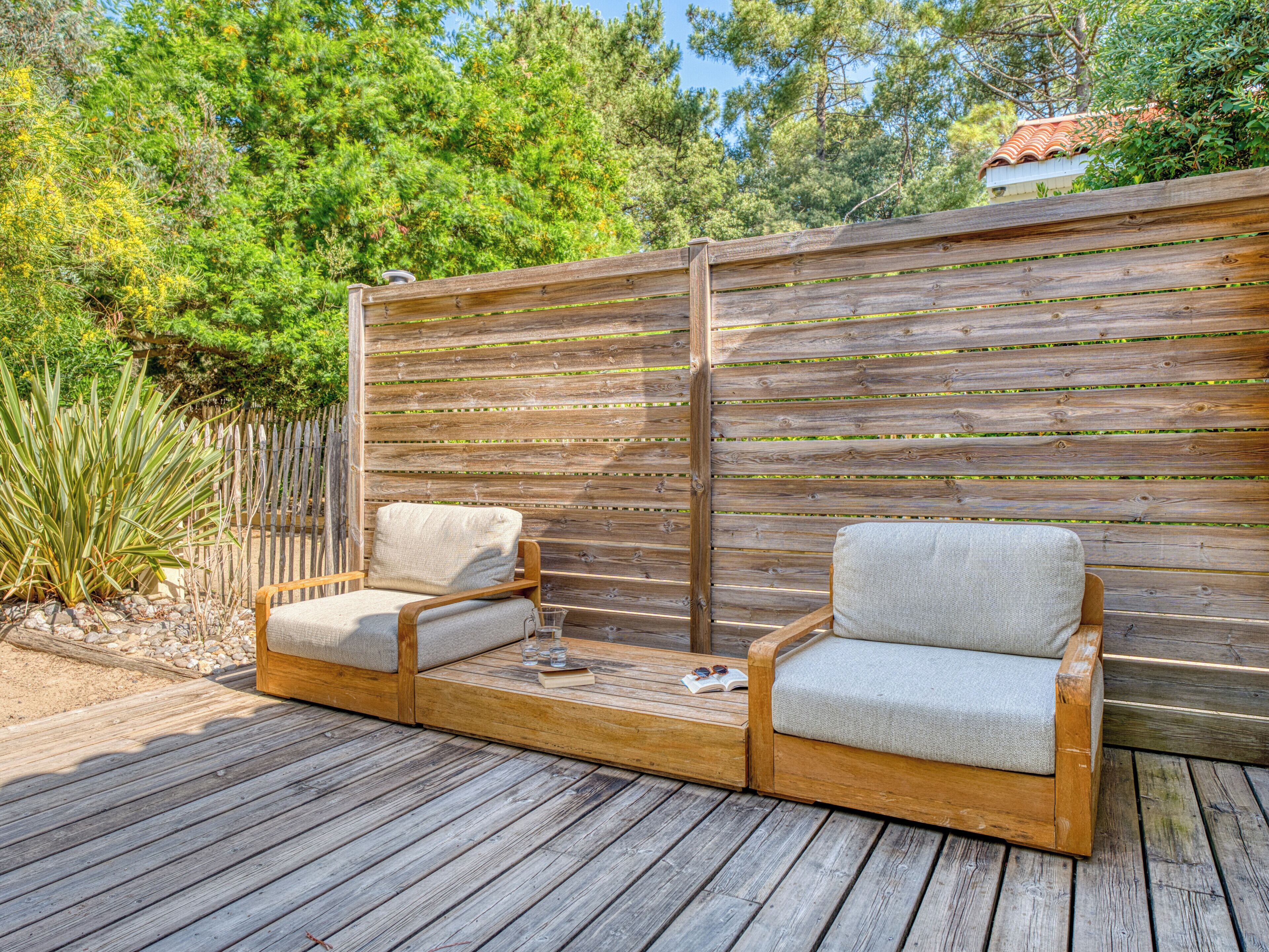 Een houten terras met twee zithoeken met kussens, een houten omheining en een achtergrond van bomen en een huis.