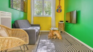 A room with a sofa, a wicker chair, a small table, a guitar, and a television on the wall. - Villa Brésil - Domaine Le Sherwood (Notre-Dame-De-Monts)