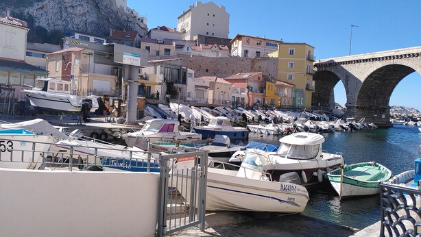 A marina with boats docked, a stone bridge, and colorful buildings in the background.