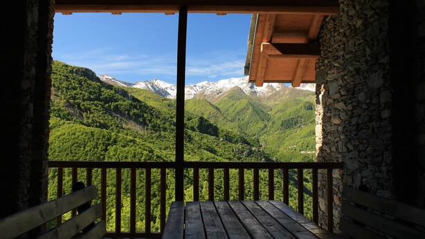 Een houten balkon met een stenen muur, met uitzicht op een bergachtig landschap met besneeuwde toppen en weelderige begroeiing.