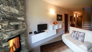 Een woonkamer met een stenen openhaard, een witte bank, een wandgemonteerde televisie en een klein wit plankje met decoratieve objecten.